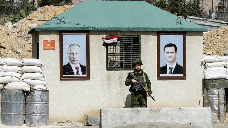 1 март куни олинган суратда Россия ҳарбий полицияси ходими Дамашқ яқинидаги ал-Вафидийн чегара пунктида Сурия президенти Башар ал-Ассад (ўнгда) ва Россия президенти Владимир Путин (чапда) портретлари ўртасида қўриқчилик постида тургани акс этган. (Louai Beshara/AFP)